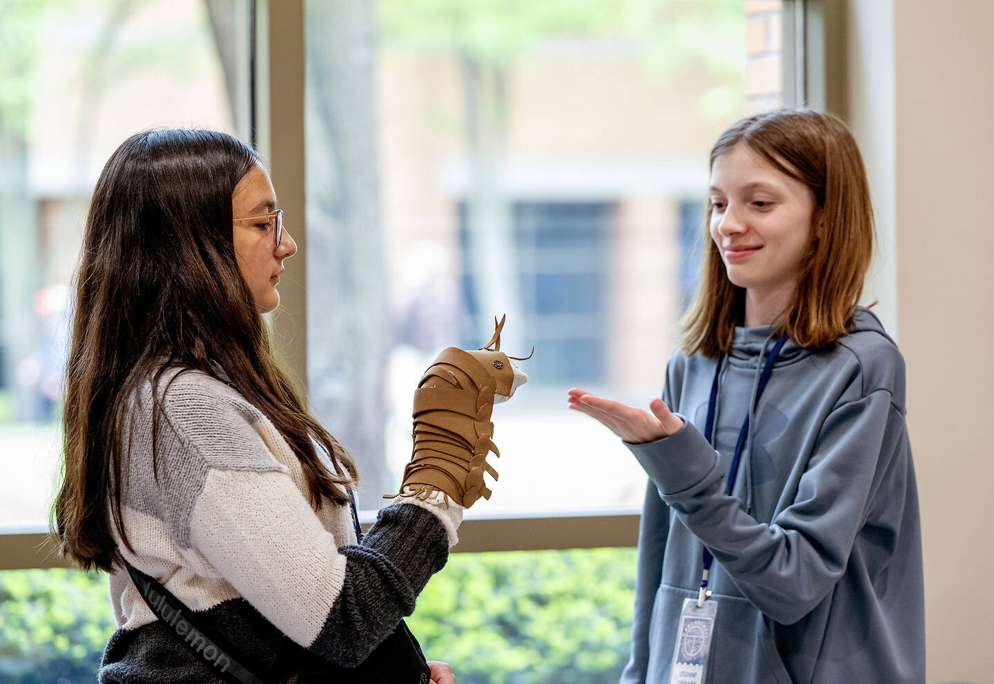 Two young people interact indoors, one holding a brown leather-like object while the other gestures with an open hand.